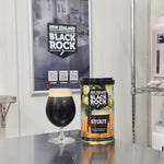 A canister of New Zealand Black Rock Stout beer stands on a stainless steel counter next to a glass filled with dark stout and a light cream head. A framed Black Rock poster hangs on the wall behind, with shelving visible at the right.